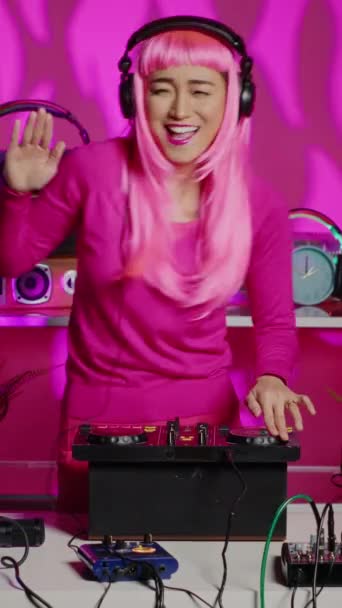 Vertical video: Smiling dj woman dancing and mixing techno sound with electronic remix using professional turntables, having fun during concert in nightclub. Asian artist standing over pink background - Záběry, video