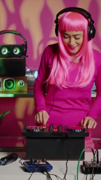 Pystysuora video: Dj nainen laittaa kuulokkeet ennen kuin aloitat musiikin sekoittamisen äänilaitteilla aikana tekno osapuoli yökerhossa. Aasian esiintyjä vaaleanpunaiset hiukset luoda musiikillista suorituskykyä mikseri - Materiaali, video