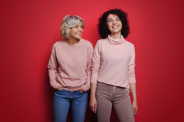 Portrait of two happy women smiling while standing over red background. Smile emotions. Looking camera. Fashion lady. Happy people positive emotions. - Photo, image