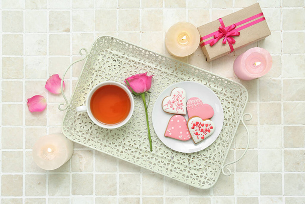 Σύνθεση με νόστιμα μπισκότα σε σχήμα καρδιάς, φλιτζάνι τσάι, κουτί δώρου και αναμμένα κεριά σε ελαφρύ πλακάκι. Γιορτή του Αγίου Βαλεντίνου - Φωτογραφία, εικόνα
