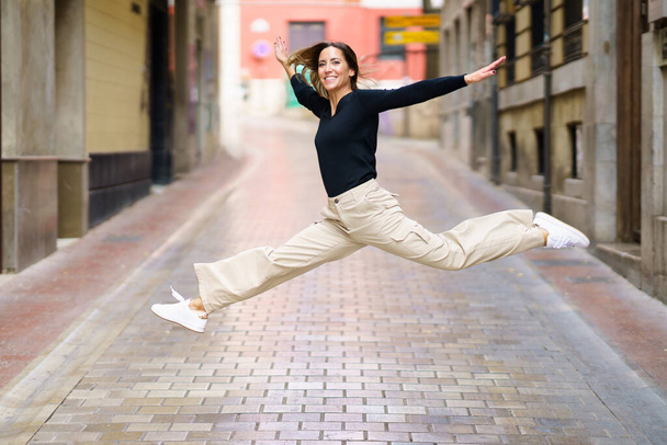 Боковой вид восхитительной молодой женщины с каштановыми волосами в повседневной одежде и кроссовках, улыбающейся и смотрящей в камеру во время прыжка по узкой асфальтированной улице - Фото, изображение