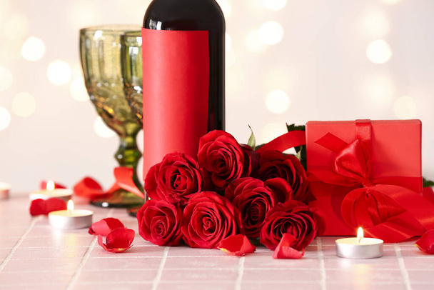 Bottle of wine, rose flowers, burning candles and glass on tile table against blurred lights. Valentine's Day celebration - Photo, image