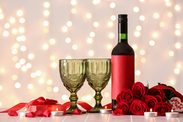 Μπουκάλι κρασί, τριαντάφυλλο λουλούδια, κεριά και ποτήρια στο τραπέζι πλακιδίων ενάντια στα θολά φώτα. Γιορτή του Αγίου Βαλεντίνου - Φωτογραφία, εικόνα