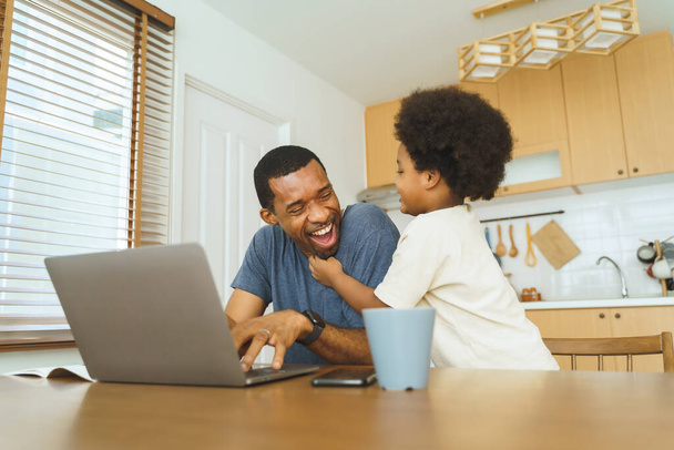 African American father working from home on laptop while his Cheerful son distracts him in kitchen. - Foto, immagini
