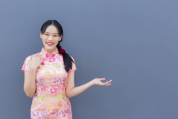 Beautiful Asian woman with long hair who wears pink Cheongsam dress in Chinese new year theme while her hand shows to present something and looks at camera smiling happily with the grey background. - Photo, Image
