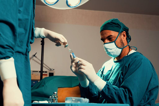 Chirurg füllen Spritze aus medizinischer Ampulle für chirurgischen Eingriff im sterilen Operationssaal mit Assistenzschwester. Arzt und medizinisches Personal in voller Schutzkleidung für die Operation bereiten Narkoseinjektionen vor - Foto, Bild