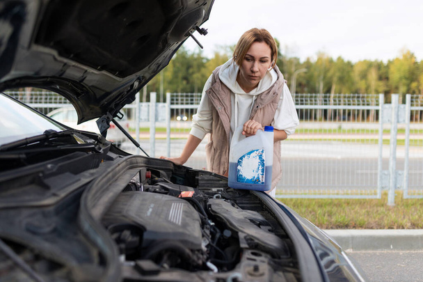 зрелая женщина заливает охлаждающую жидкость в двигатель сломанной машины. - Фото, изображение