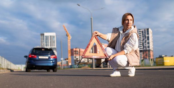 dojrzała kobieta umieszcza awaryjny znak stopu przed zepsutym samochodem na drodze. - Zdjęcie, obraz