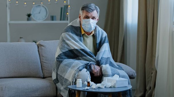 Enfermo abuelo trastornado en máscara médica sentado en sofá caucásico anciano envuelto en manta se siente escalofríos maduro anciano enfermo mirando los síntomas de la cámara covid 19 epidemia coronavirus brote gripe concepto - Foto, Imagen