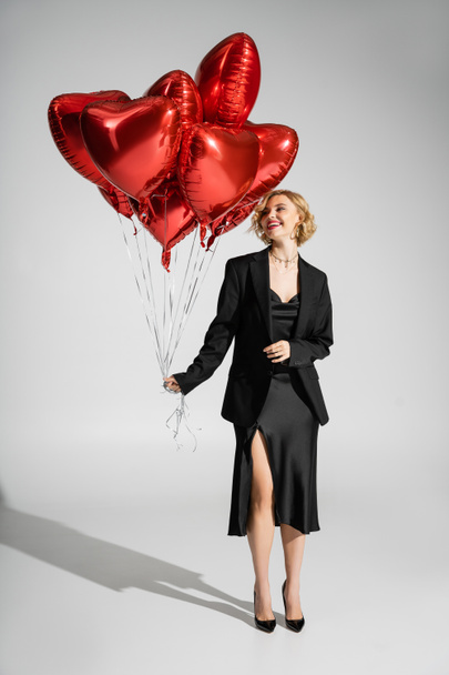 full length of cheerful woman in black slip dress and blazer looking at red heart-shaped balloons on grey  - Fotografie, Obrázek