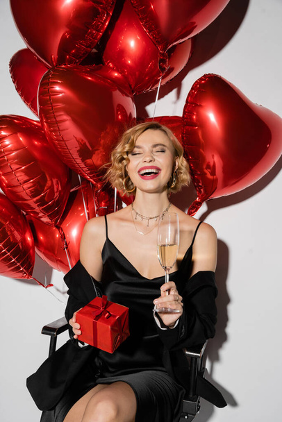 happy woman in black slip dress holding glass of champagne and present while sitting near red balloons on grey  - Фото, зображення