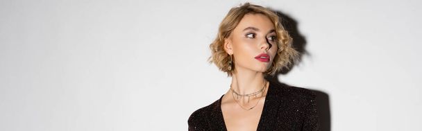 blonde young woman in golden hoop earrings and silver necklace looking away on grey background, banner  - Foto, imagen