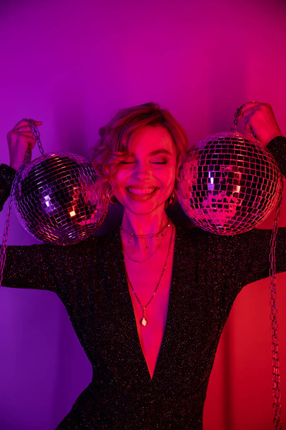 cheerful young woman with blonde hair standing in black dress and holding chains with disco balls on purple and pink  - Photo, image