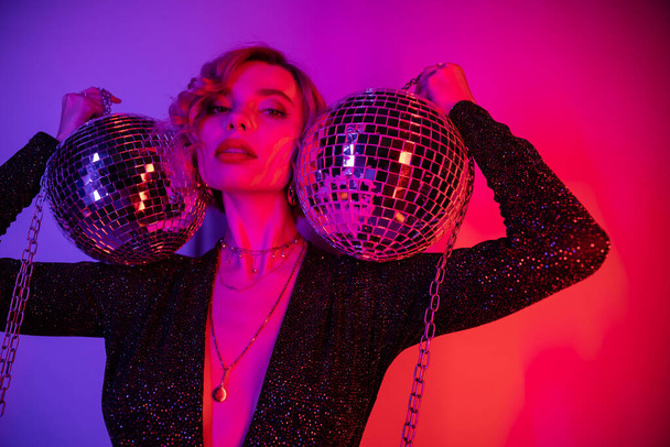young woman with blonde hair standing in black dress and holding chains with disco balls on purple and pink  - Fotografie, Obrázek