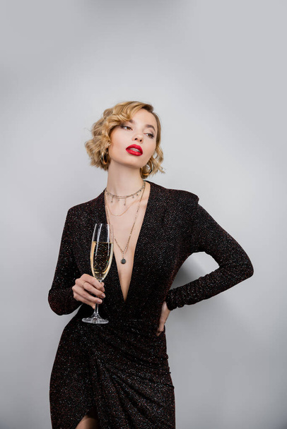 young woman with blonde curly hair and red lips posing with hand on hip while holding glass of champagne on grey  - Foto, Imagem