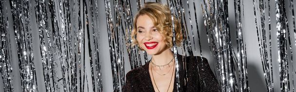 positive young woman with red lips standing in black dress near shiny tinsel curtain on grey, banner  - Foto, Imagen