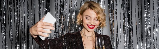 iloinen nainen ottaa selfie pitäen lasi samppanjaa lähellä tinsel verho harmaa, banneri  - Valokuva, kuva
