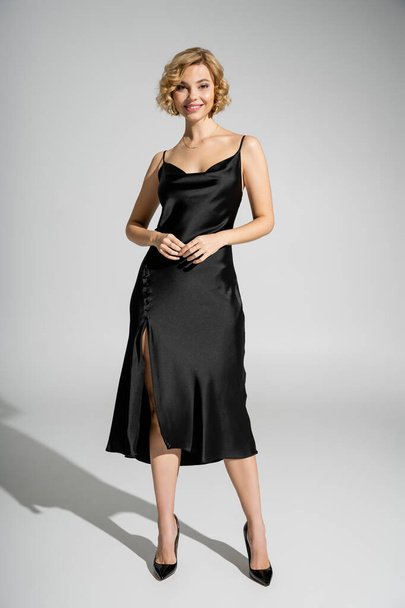 full length of cheerful and blonde woman in elegant slip dress standing on grey  - Photo, image
