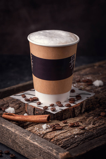 Картонный стакан с кофе на деревянной доске, кофейные зерна разбросаны по доске и лежит корица палка - Фото, изображение