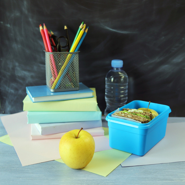Обідня коробка з бутербродом, пляшка води, яблуко, книги та канцелярські товари на дерев'яному столі, фон з крейди, назад до школи
  - Фото, зображення