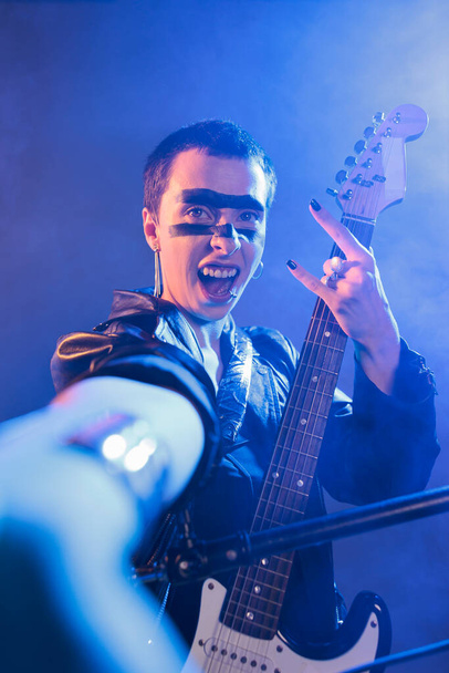 Crazy artist playing bass guitar in studio with smoke, showing rock sign with fingers and acting stylish. Cool ecstatic rocker creating live performance with heavy metal and punk music. - Photo, image