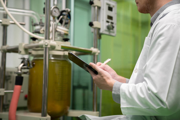 Closeup apothecary scientist using a clipboard and pen to record information from a CBD oil extractor and a scientific machine used to create medicinal cannabis products. - Photo, Image