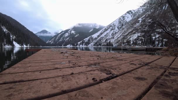 A guy walks along a pier on a mountain lake. Dark water like a mirror reflects snowy mountains, forest and clouds. Yellow sunset. A place for meditation and enjoyment. Epic. Lake Kolsai, Kazakhstan. - Footage, Video