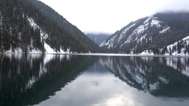 Гірське озеро в лісі з дзеркальною водою. Дерев'яний пірс з білими човнами. Вода віддзеркалює пейзаж хмарного неба, засніжених гір і вершин, хвойних ярів. Озеро Колсай, Казахстан - Кадри, відео