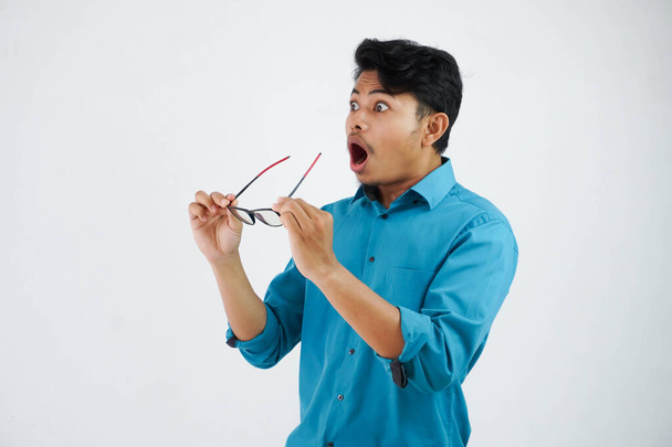 Překvapený nebo Shock hezký asijský muž s brýlemi v modré košili otevření úst a rukou drží brýle na izolovaném bílém pozadí - Fotografie, Obrázek