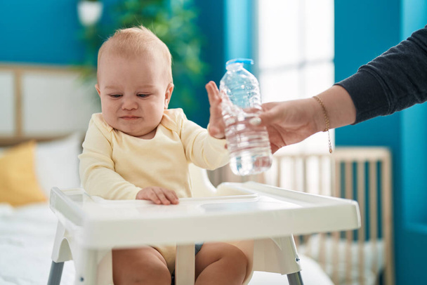 Adorable bebé caucásico sentado en trona rechazando botella de agua en el dormitorio - Foto, Imagen