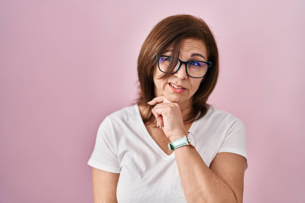 Mujer hispana de mediana edad de pie sobre fondo rosa pensando preocupado por una pregunta, preocupado y nervioso con la mano en la barbilla  - Foto, Imagen