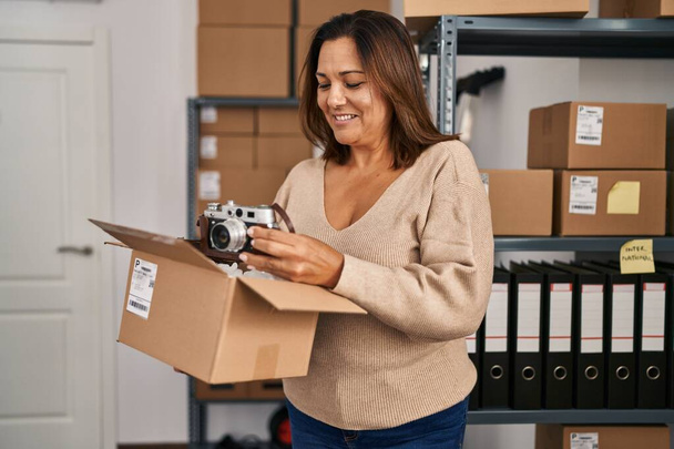 Middle age hispanic woman ecommerce business worker unpacking camera at office - Фото, изображение