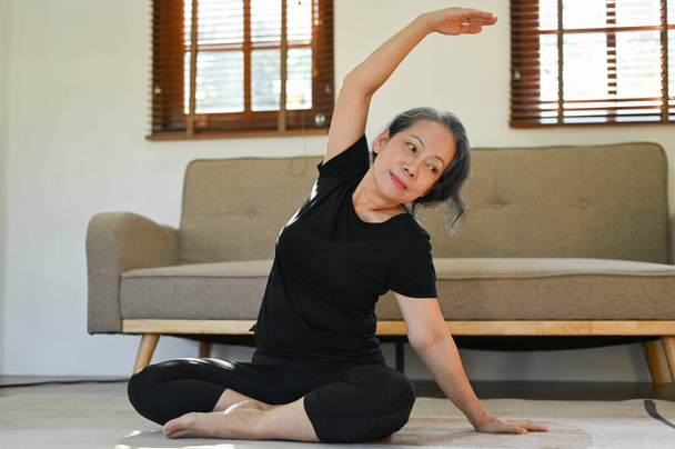 Terve ja onnellinen 60-luvulla eläkkeellä oleva aasialainen nainen harjoitusvaatteissa harjoitellen joogaa olohuoneessaan. - Valokuva, kuva
