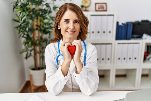 Mulher hispânica de meia idade vestindo uniforme médico segurando coração na clínica - Foto, Imagem