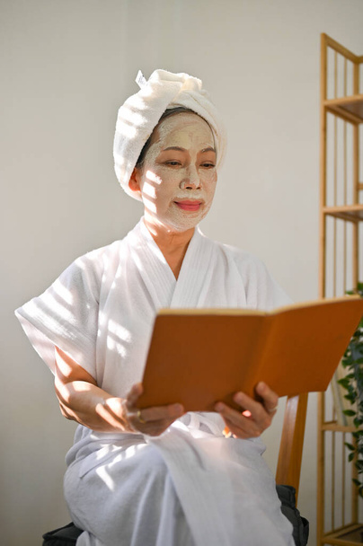 Ελκυστική και χαρούμενη ώριμη Ασιάτισσα με μπουρνούζι διαβάζει ένα βιβλίο ενώ εφαρμόζει μάσκα προσώπου από πηλό στο πρόσωπό της. Έννοια ημέρας περιποίησης ομορφιάς - Φωτογραφία, εικόνα