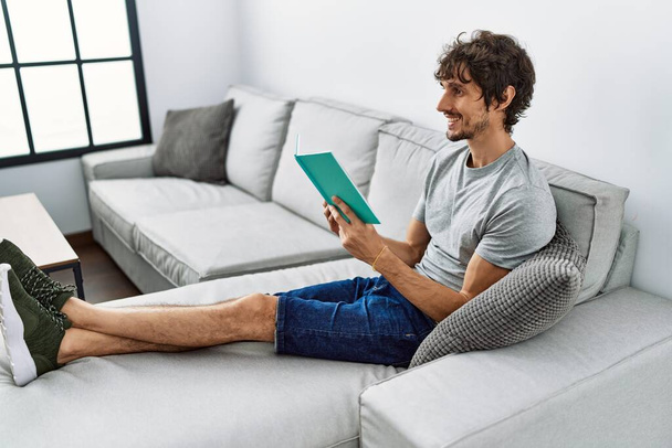 Young hispanic man smiling confident reading book at home - 写真・画像