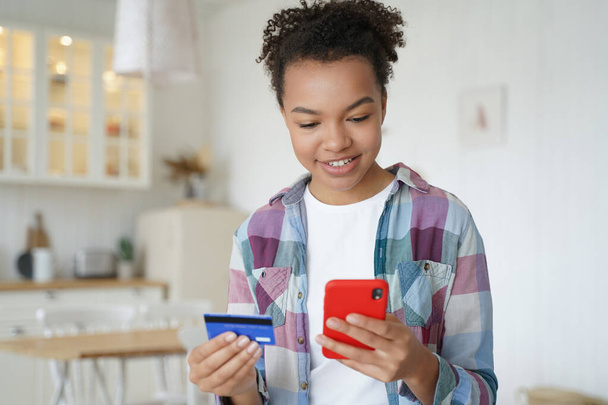 Happy mixed race teen girl holding credit card, shopping on smartphone. Smiling modern young female paying for purchases on internet using mobile bank app, online banking services, standing at home. - Foto, imagen