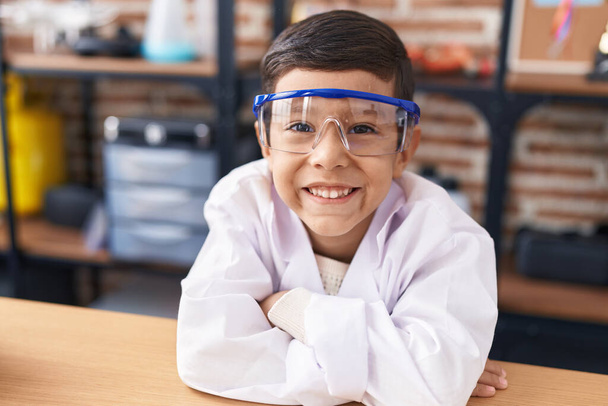 Adorable niño hispano estudiante apoyado en la mesa con los brazos cruzados gesto en el aula de laboratorio - Foto, Imagen