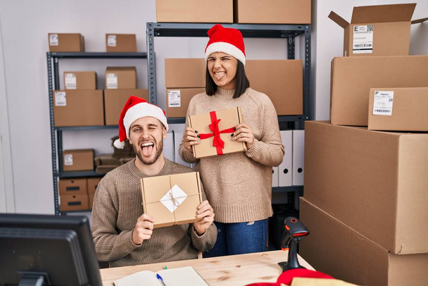 Zwei junge Leute, die zu Weihnachten im E-Commerce eines Kleinunternehmens arbeiten, strecken mit lustiger Miene die Zunge heraus.  - Foto, Bild