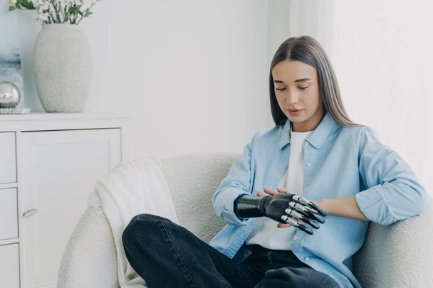 Handicapped girl looks and touches her bionic arm. Young pensive european woman with cyber hand at home. Modern bionic prosthesis. Futuristic technology of artificial limbs. - Zdjęcie, obraz
