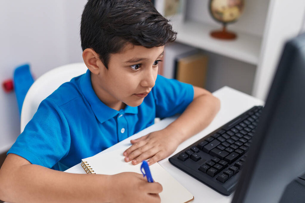 Adorable hispanic boy student using computer writing on notebook at classroom - Zdjęcie, obraz