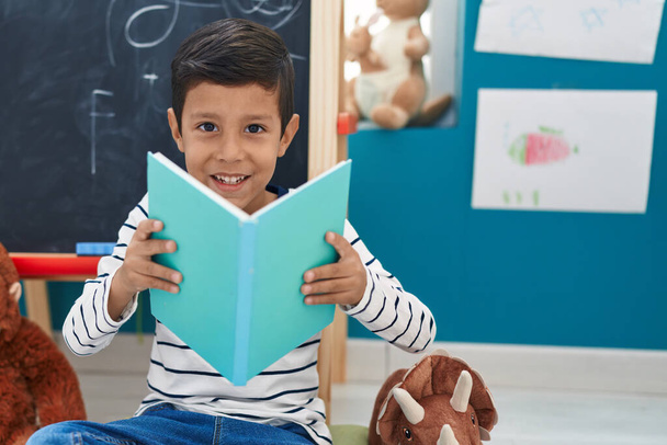 Adorable hispanic boy student smiling confident reading book at kindergarten - Photo, Image