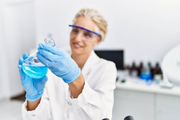 Middle age blonde woman wearing scientist uniform holding test tube at laboratory - Foto, immagini
