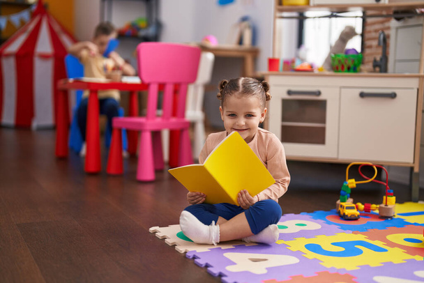 Adorable estudiante hispana sentada en el libro de lectura de piso en el jardín de infantes - Foto, Imagen