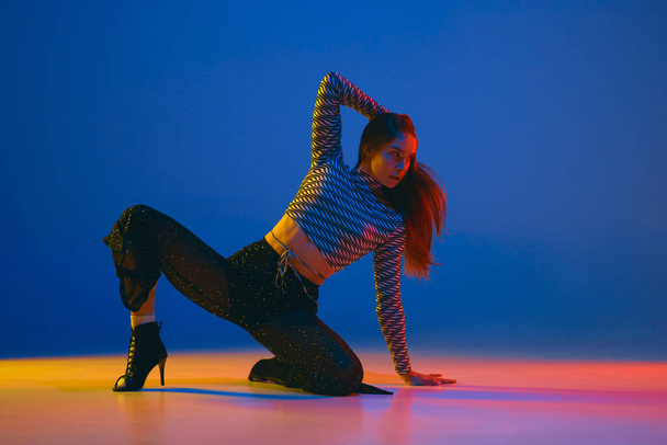 Passion. Portrait of young girl dancing high heel dance in stylish clothes over blue background in neon light. Concept of dance lifestyle, modern style, contemporary, youth culture, self-expression - Photo, Image
