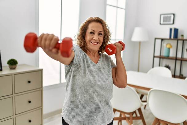 Middle age caucasian woman smiling confident training using dumbbells at home - Fotografie, Obrázek