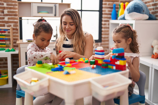 Вчителька з дівчатами грає з будівельними блоками, сидячи на столі в дитячому саду
 - Фото, зображення
