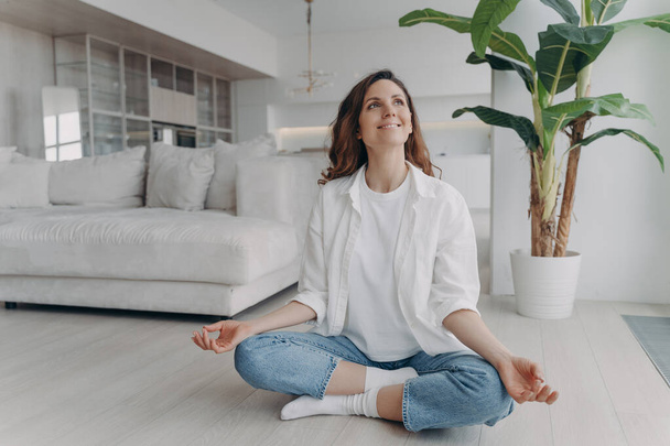 Peace of mind and mental health concept. Young european woman is practicing yoga on floor and smiling. Meditation, consciousness and relaxation at home. Posture exercise, lotus asana. - Photo, Image