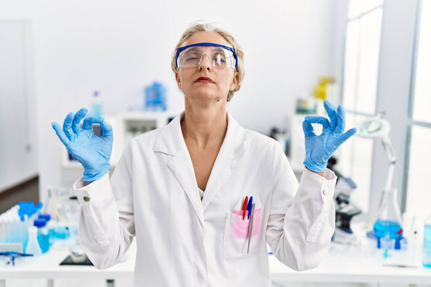 Светловолосая женщина средних лет, работающая в лаборатории, расслабляется и улыбается с закрытыми глазами, делая медитационный жест пальцами. Концепция йоги.  - Фото, изображение
