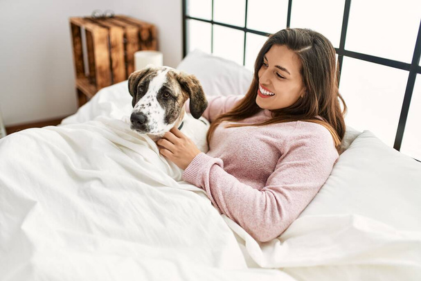 Mujer joven sonriendo confiado abrazando perro sentado en la cama en el dormitorio - Foto, Imagen